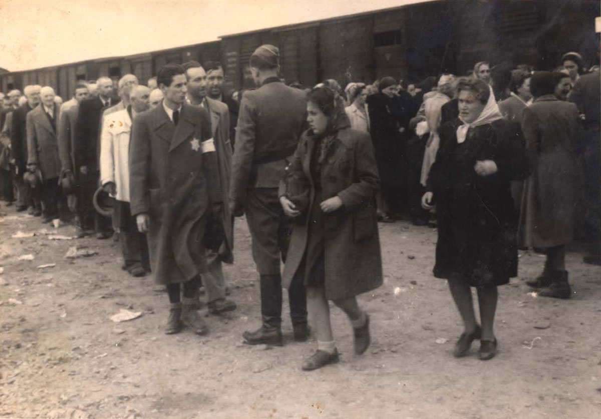 Die Reihe der jüdischen Männer auf der linken Seite bewegt sich auf einen SS-Mann zu. Rechts dahinter findet die Selektion der jüdischen Frauen statt. Die beiden Frauen im Vordergrund wurden anscheinend gerade als Zwangsarbeiterinnen selektiert